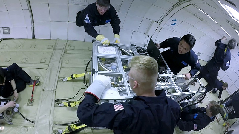 A group of astronauts in a space station work together around a metal container filled with equipment. They are all dressed in dark uniforms.