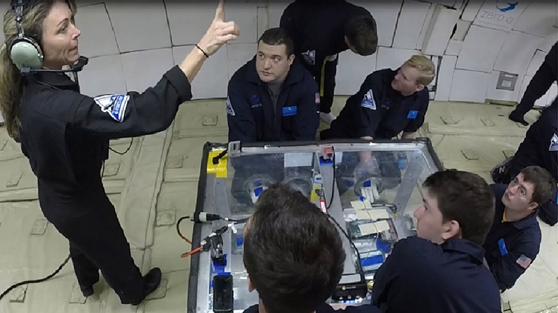 A group of people in black uniforms gather around a table with equipment, as a woman with a headset points upwards while speaking. The setting appears to be zero-gravity, suggested by the surroundings.