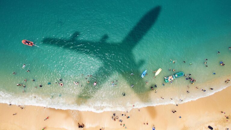 Aerial view of a beach with swimmers and kayakers in the ocean. The shadow of a large airplane is visible on the water surface.