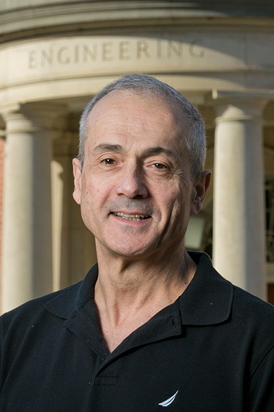 A man with short gray hair wearing a black polo shirt stands outside an engineering building with columns.