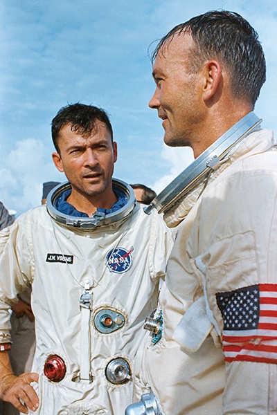 Two astronauts in spacesuits with NASA patches and American flag armbands standing outside, one facing the camera while the other faces him.