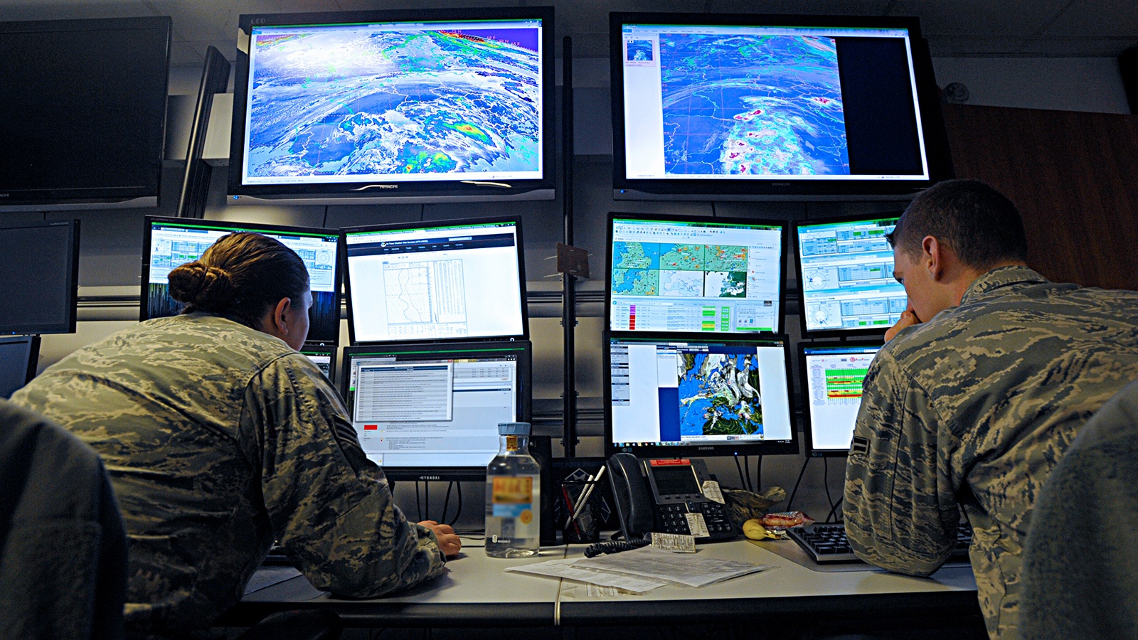 Two military personnel monitor multiple screens displaying weather patterns and maps in a control room.