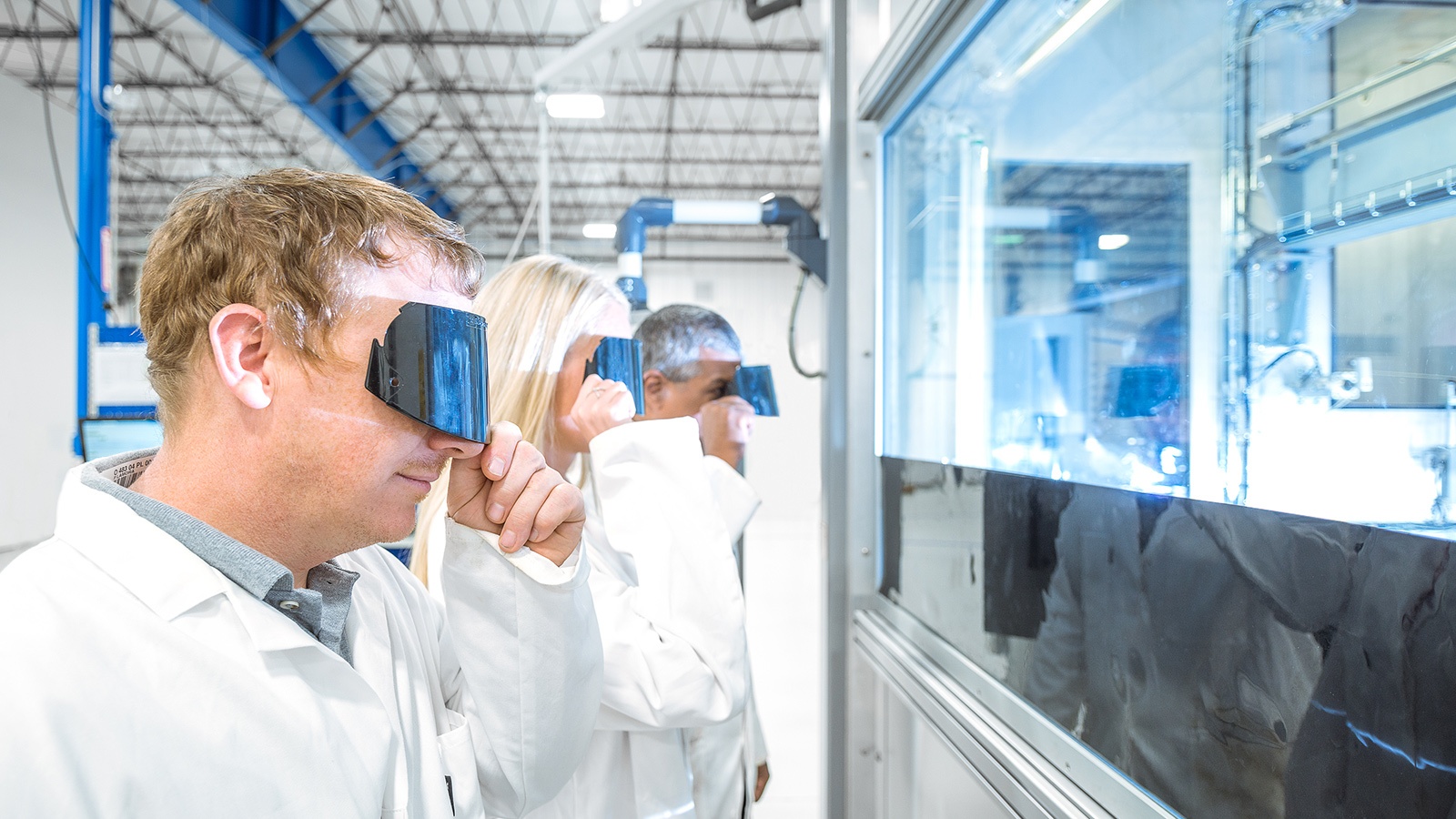 Three people in lab coats observe a bright light source through protective lenses in an industrial facility.