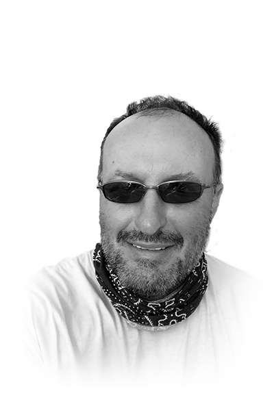 A man wearing sunglasses, a white shirt, and a patterned scarf around his neck smiles at the camera.