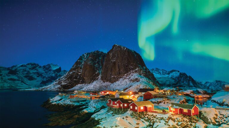 Small red cabins sit along a snowy shoreline beneath the Northern Lights, with a large rocky mountain in the background and a starry night sky above.