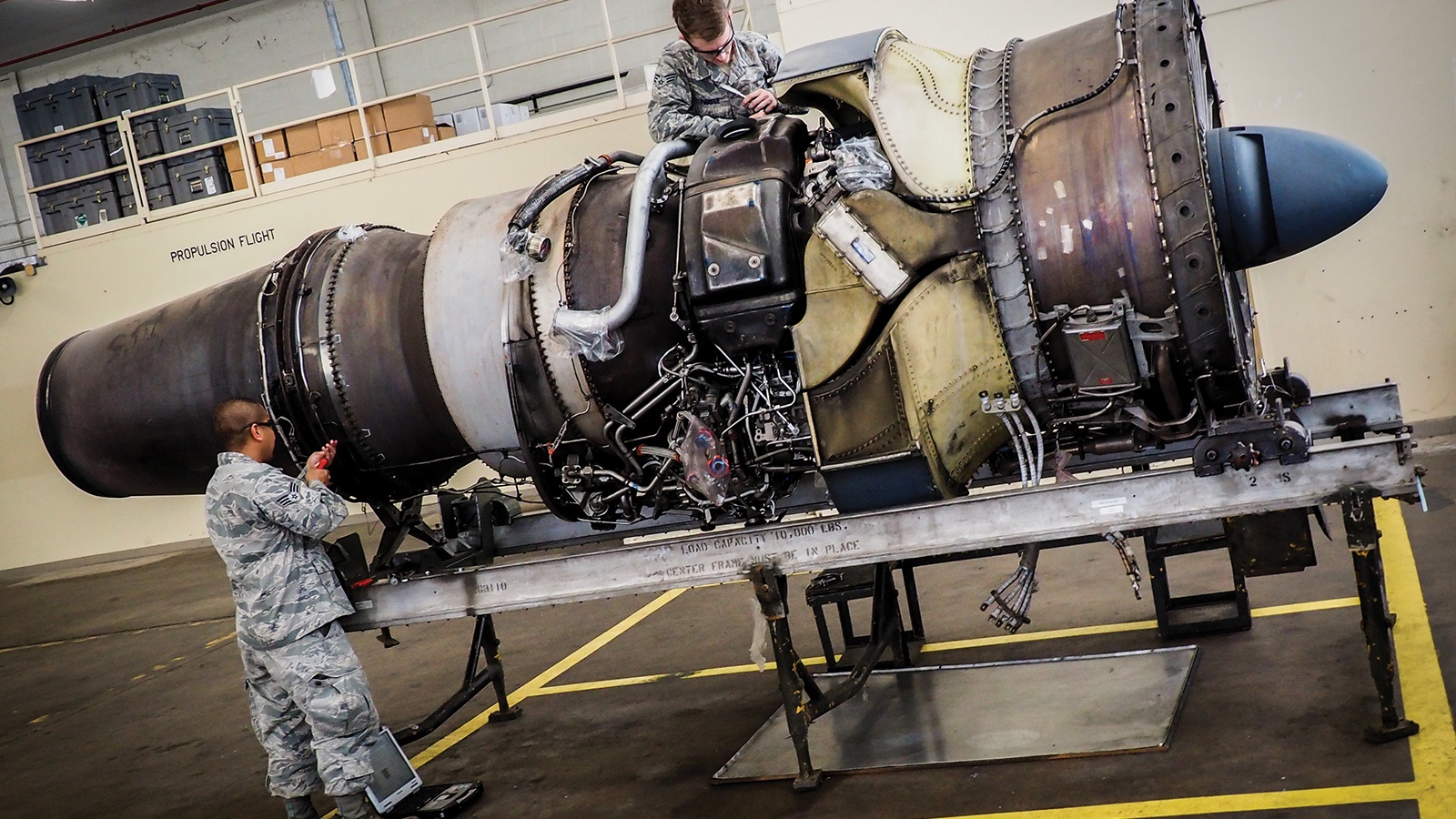 Two people in military uniforms work on an aircraft engine mounted on a stand inside a maintenance facility.