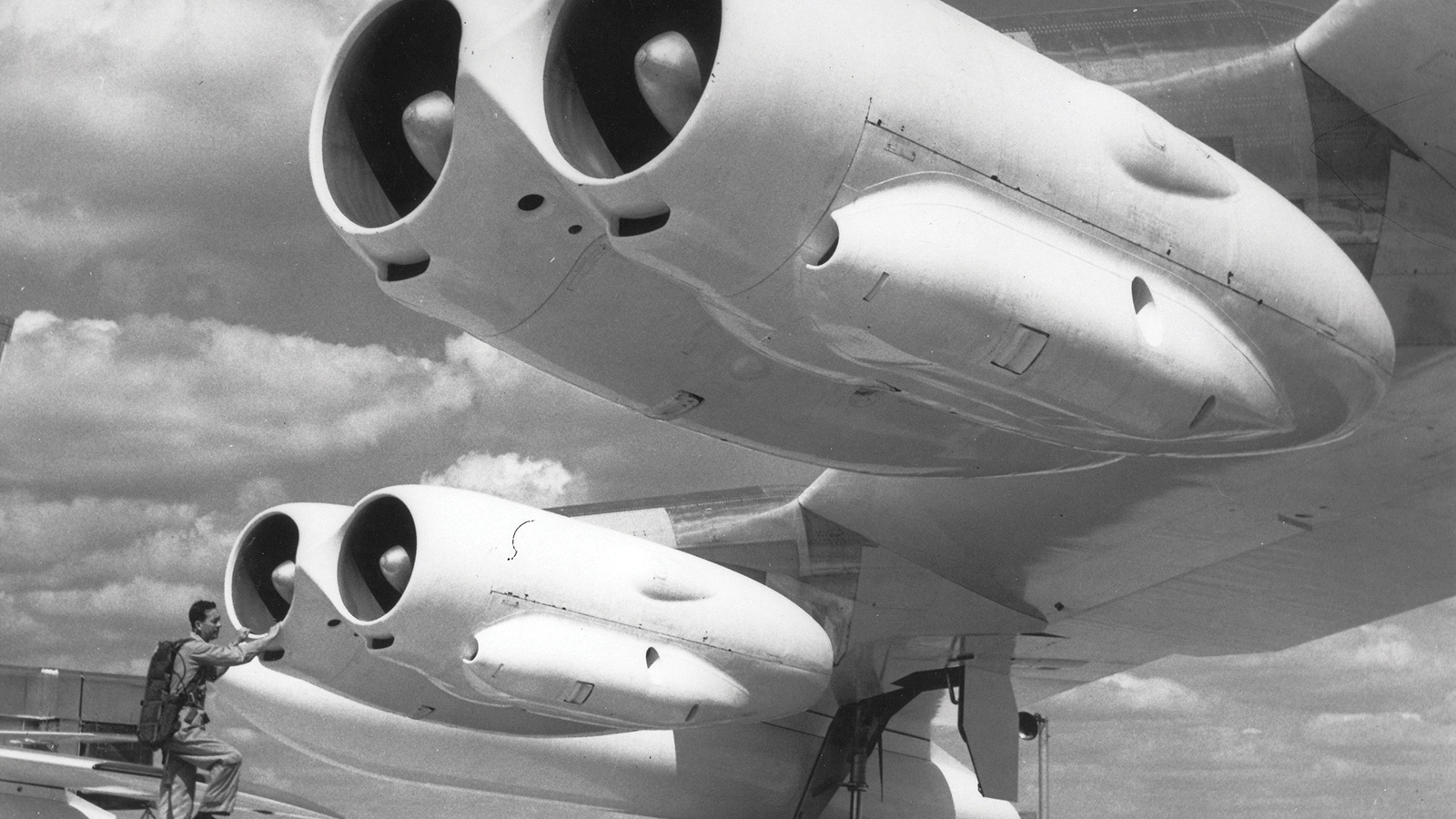 Black and white photo of a large aircraft's underbelly showing its three sizable jet engines. A person stands near the engines, seemingly inspecting them, against a backdrop of a partly cloudy sky.