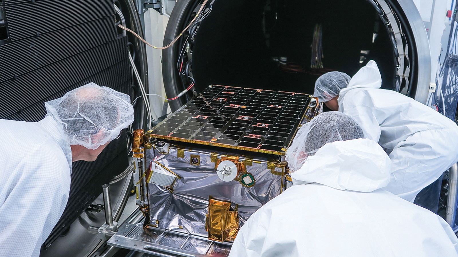 Engineers in white lab coats and hairnets examine a satellite or spacecraft being placed into a testing chamber.