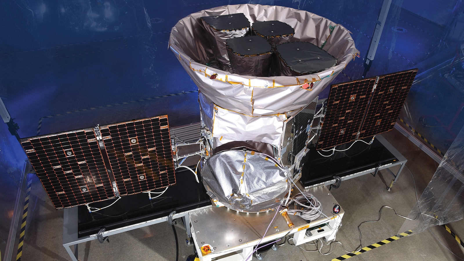 A satellite with solar panels and a large, cone-shaped structure in a lab setting, surrounded by protective materials and equipment.