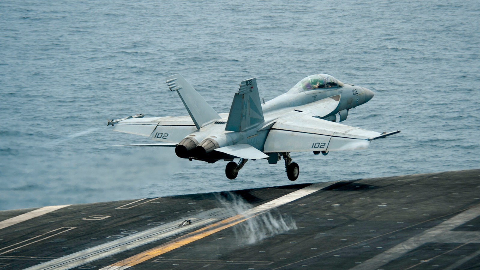 A fighter jet takes off from an aircraft carrier over the ocean, with its landing gear retracted and water sprays visible from the deck.