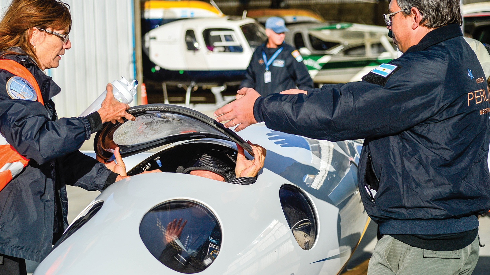 A person is preparing to enter a small aircraft cockpit, assisted by two individuals. Several airplanes are visible in the background.