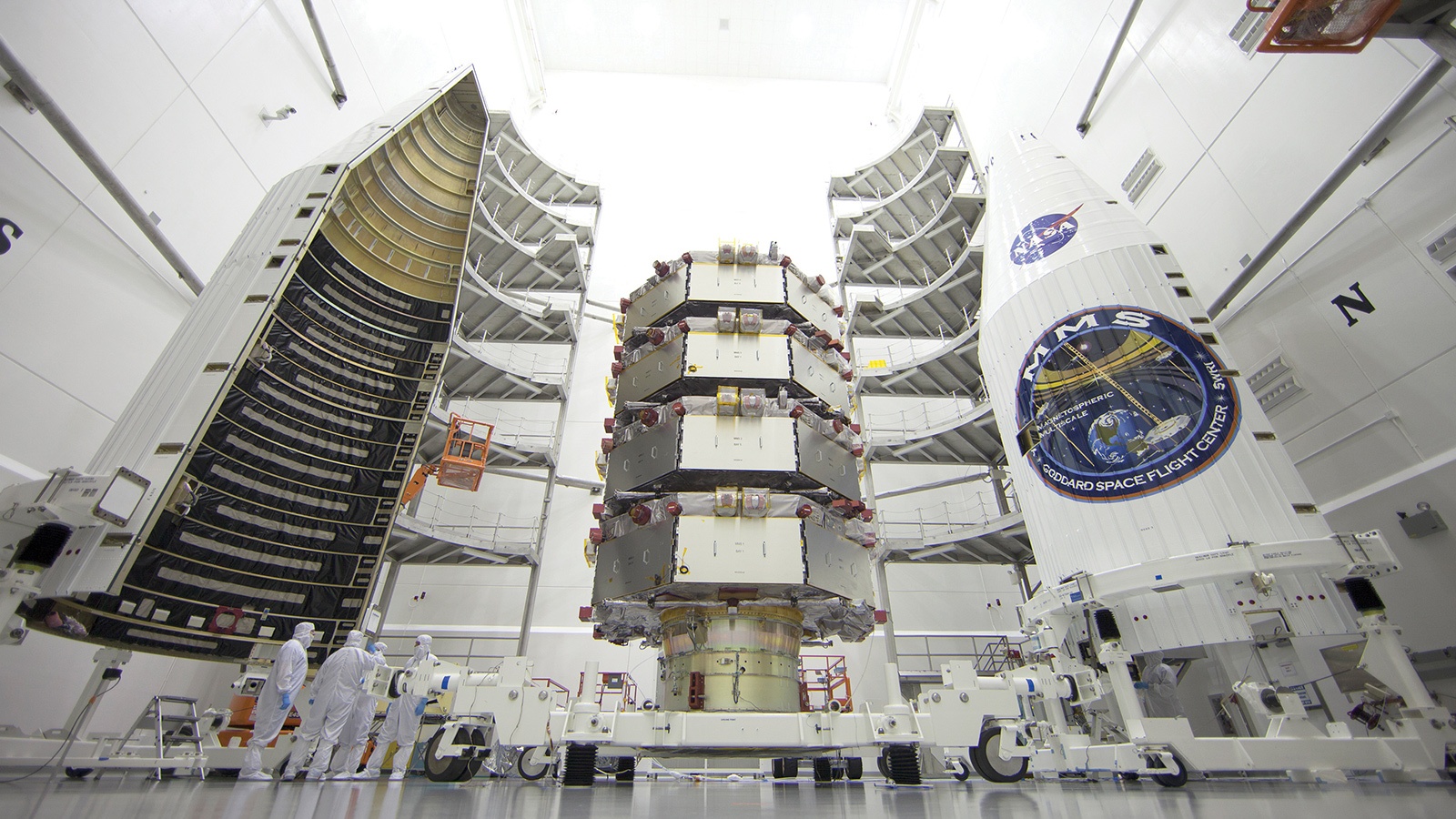 Technicians in a cleanroom work on a satellite payload as it is being enclosed in a rocket fairing, with part of the fairing open on the left and part closed on the right.