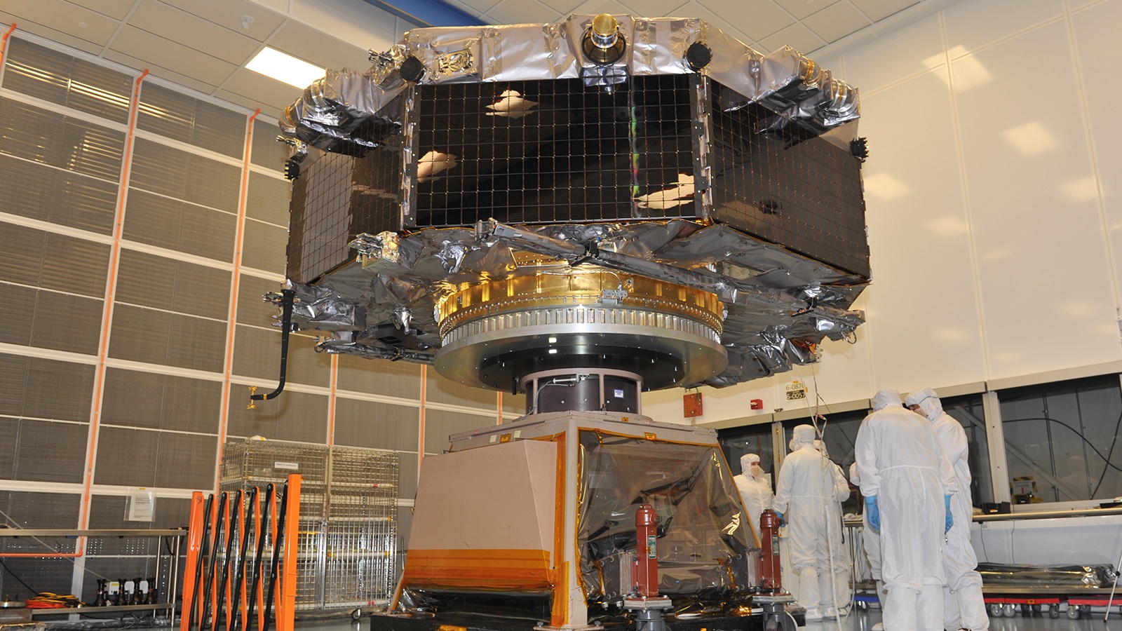 A large spacecraft is being assembled in a cleanroom by several technicians wearing white protective suits.
