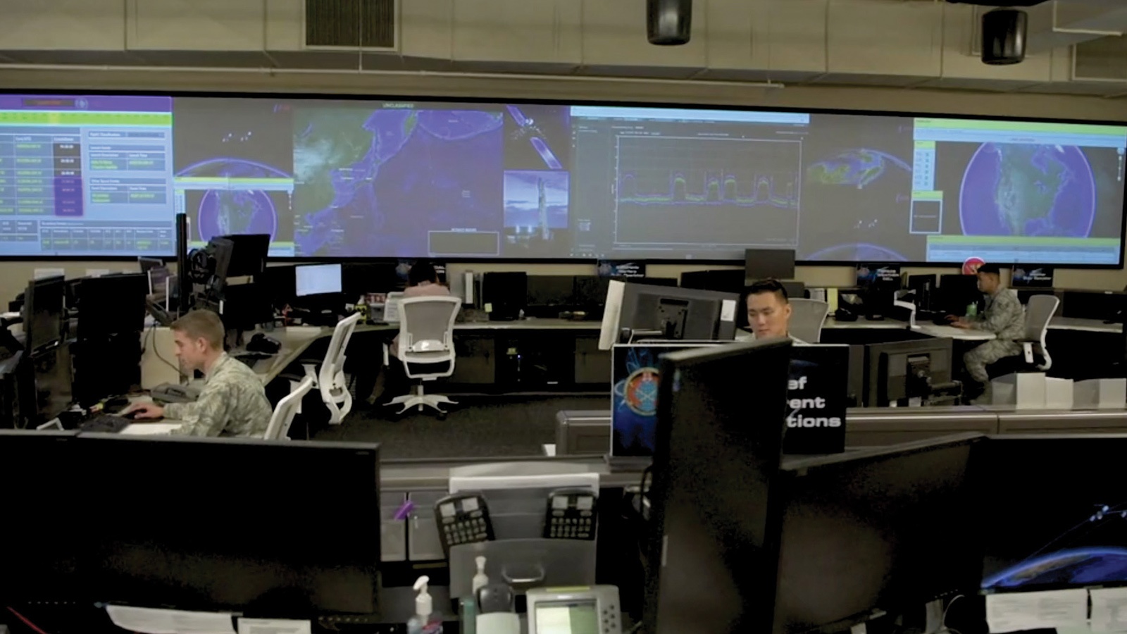 A high-tech operations center with several personnel working at computer stations. Large screens on the wall display maps and data.