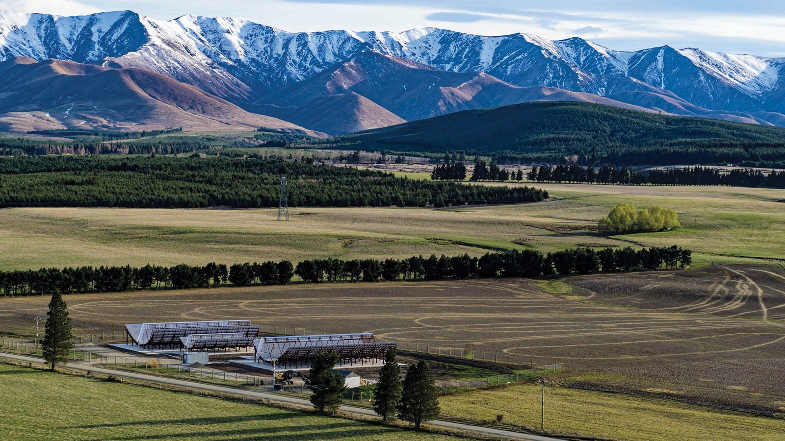 Aerial view of green fields with two structures and rows of trees in the foreground, set against a backdrop of rolling hills and snow-capped mountains.