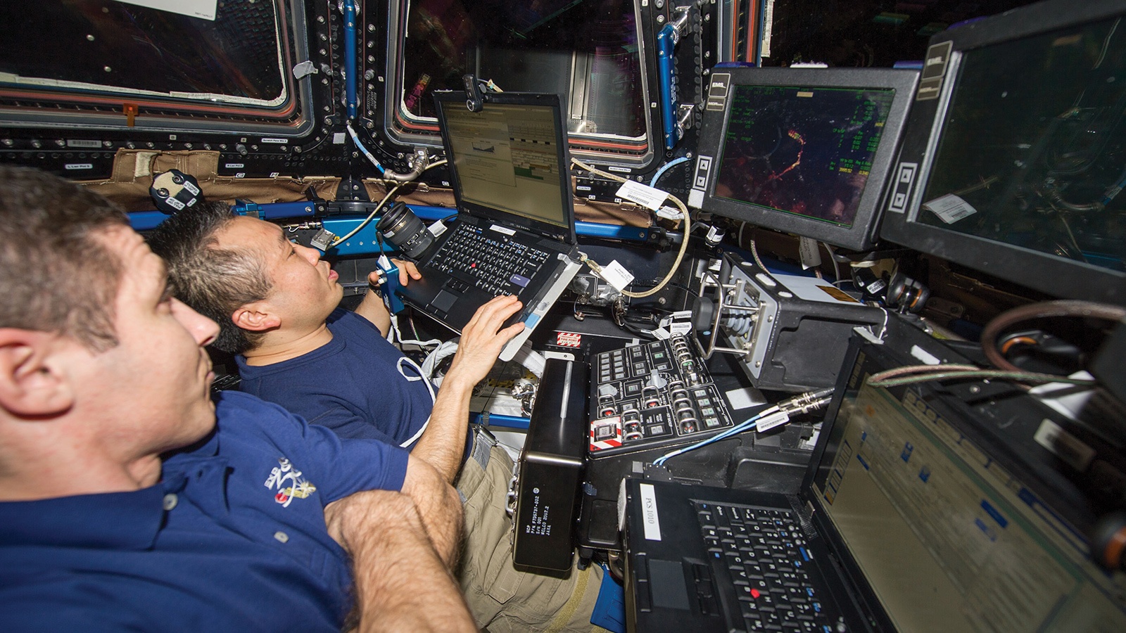 Two astronauts operate laptops and equipment inside the International Space Station, surrounded by various scientific instruments and monitors displaying data.