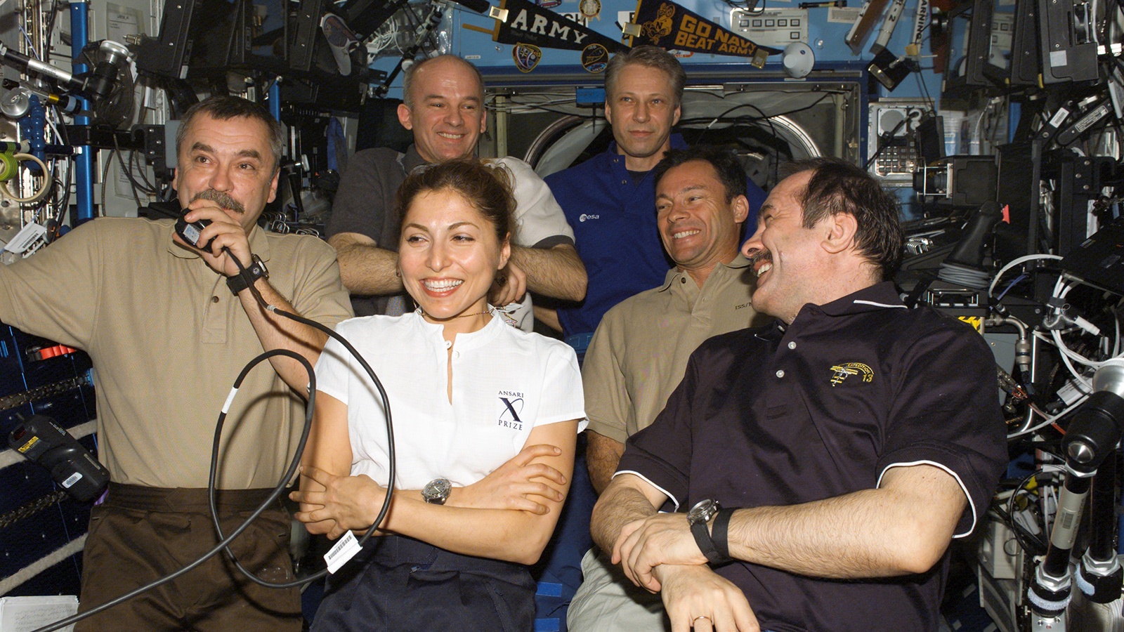 Group of six astronauts smiling and posing together inside the International Space Station, surrounded by equipment and controls.