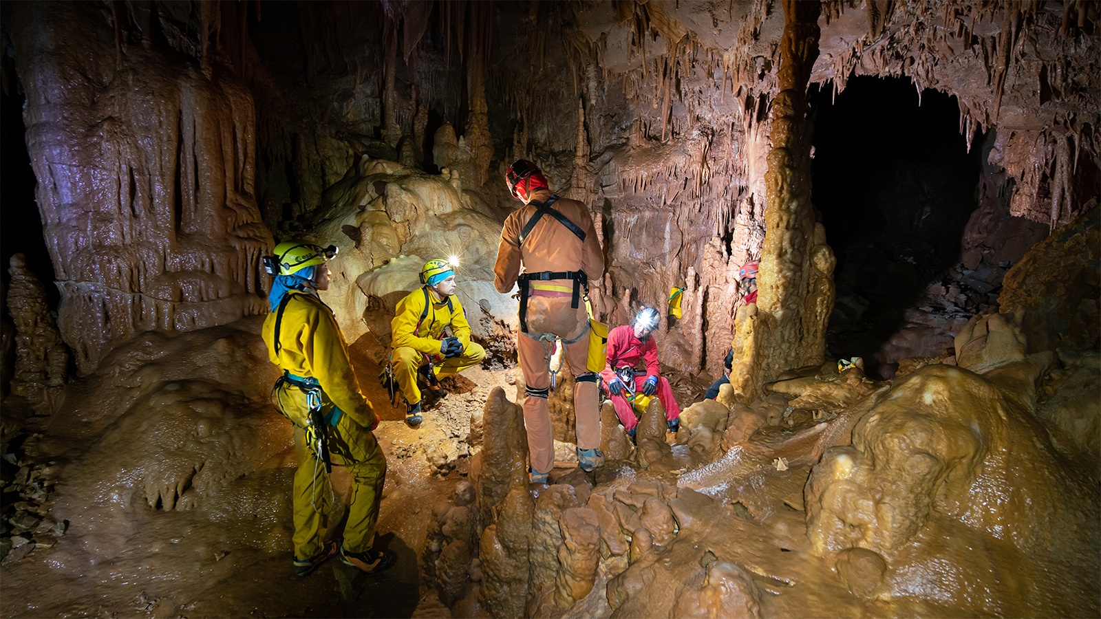 A group of cavers in protective gear gather inside a cave, examining rock formations. The cave's interior is illuminated by their headlamps, revealing stalactites and stalagmites.