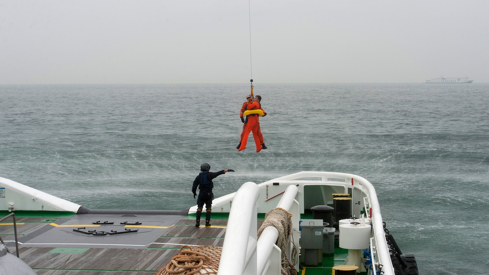 A person in an orange suit is being hoisted off a ship's deck by a cable and harness over a body of water, with another person on deck guiding the operation.