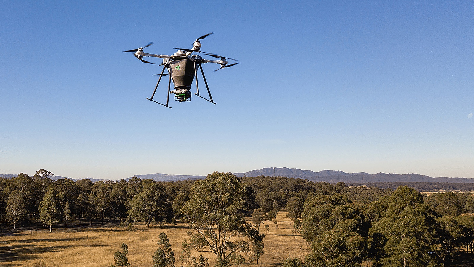 A drone flies in the sky above a rural landscape with trees and mountains in the background.