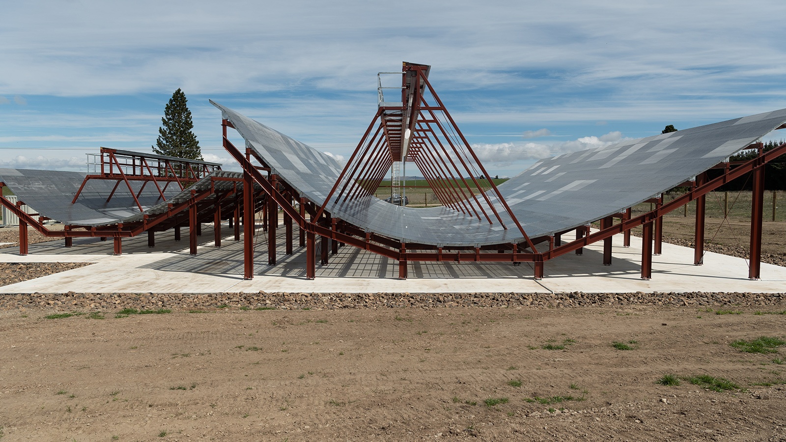 A large, outdoor scientific structure composed of wave-shaped metal frames and panels built on a concrete base, situated in a rural area with a cloudy sky overhead.