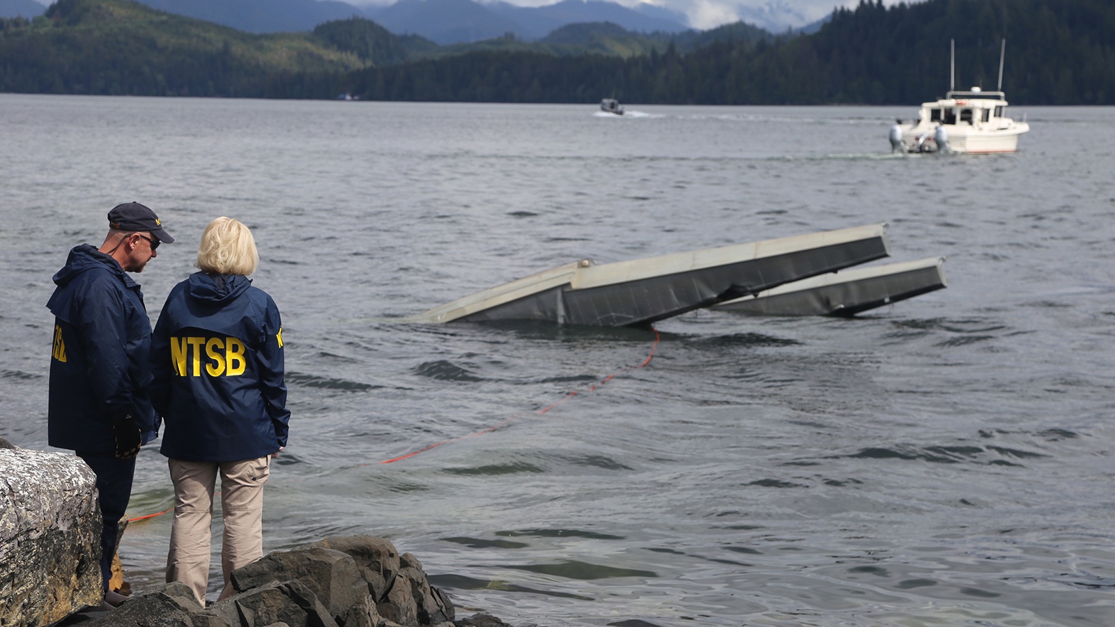 Two NTSB personnel stand on a rocky shore observing a partially submerged wreckage in a body of water with boats in the background.