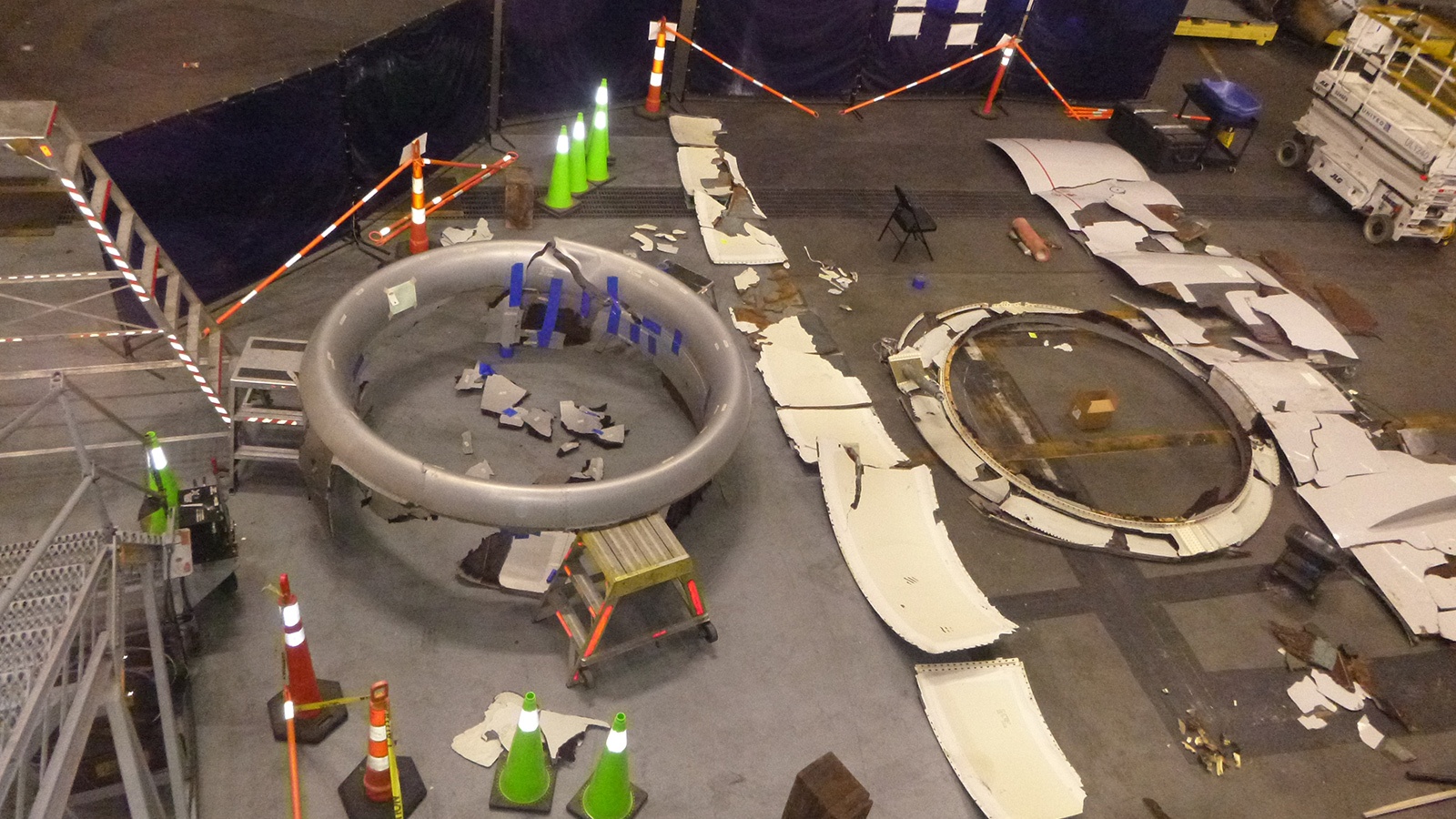 An aerial view of a work area featuring disassembled aircraft parts, including large circular components on the ground, surrounded by traffic cones, barriers, and scattered debris.