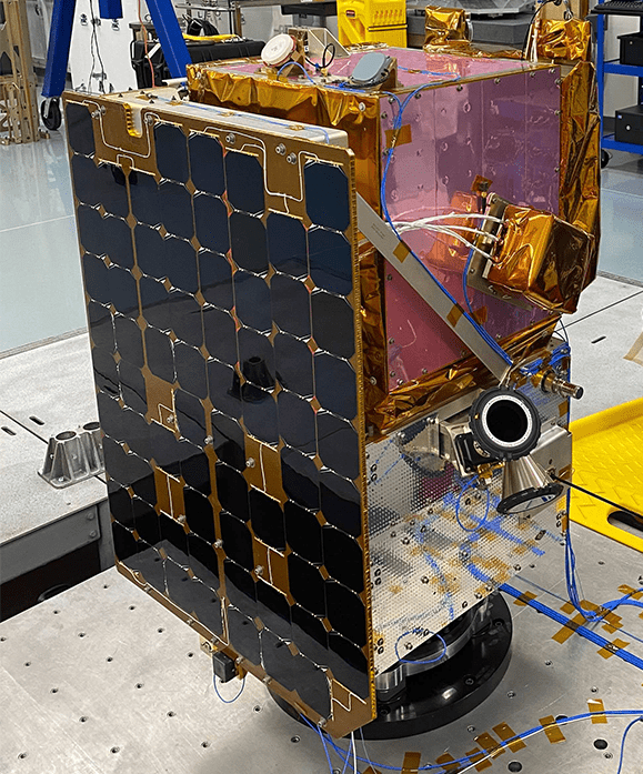 A spacecraft with solar panels and various instruments, partially wrapped in gold foil, is situated in a lab environment.