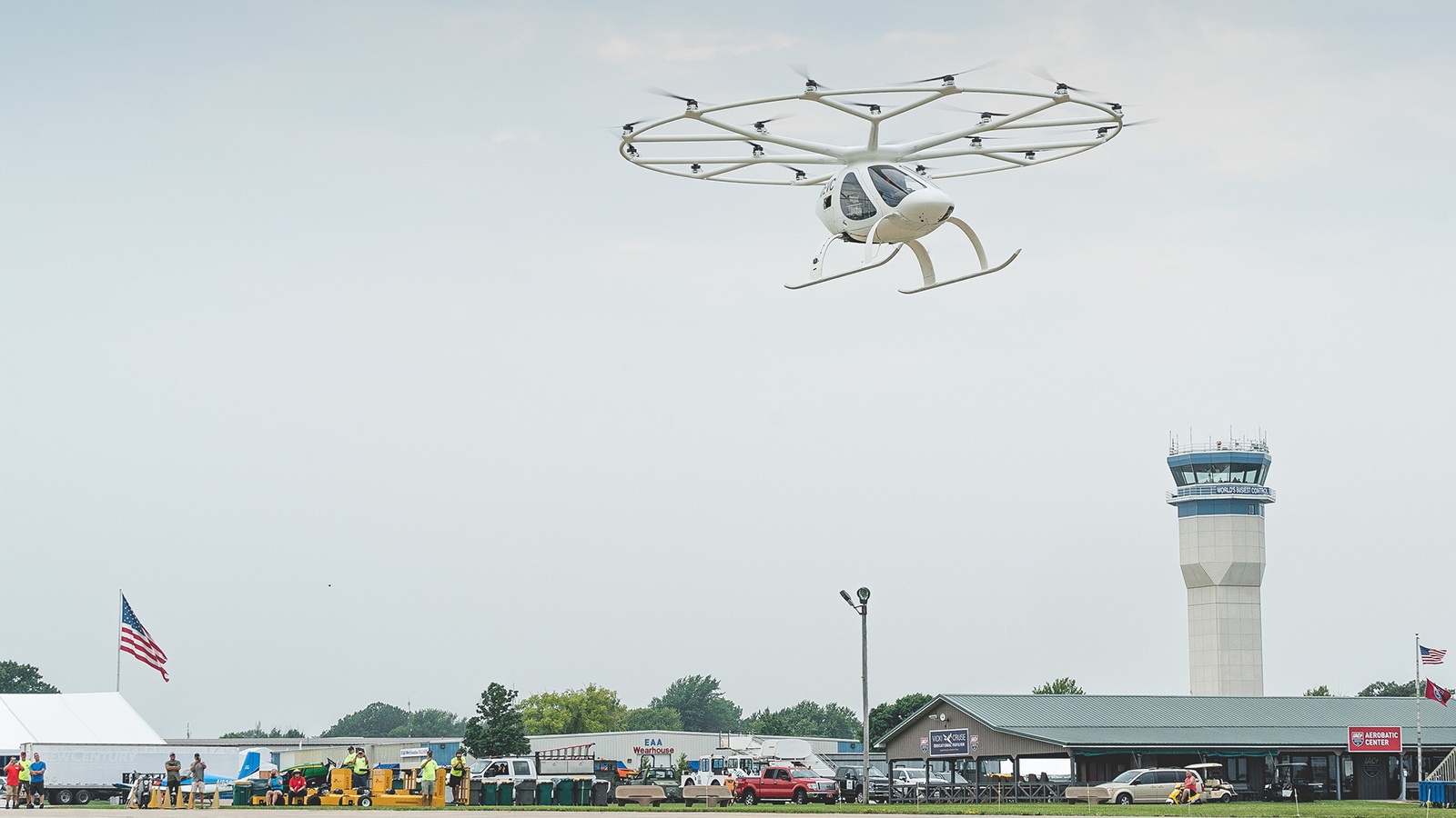 A white multicopter aircraft flies above an airfield with a control tower, small buildings, an American flag, and several vehicles on the ground beneath a foggy sky.