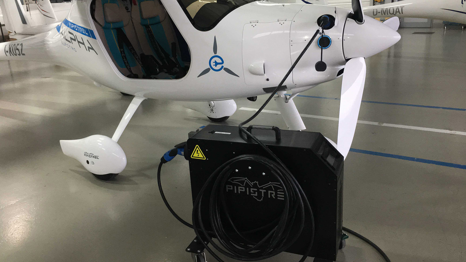 A small white electric aircraft is tethered to a black charging unit labeled 