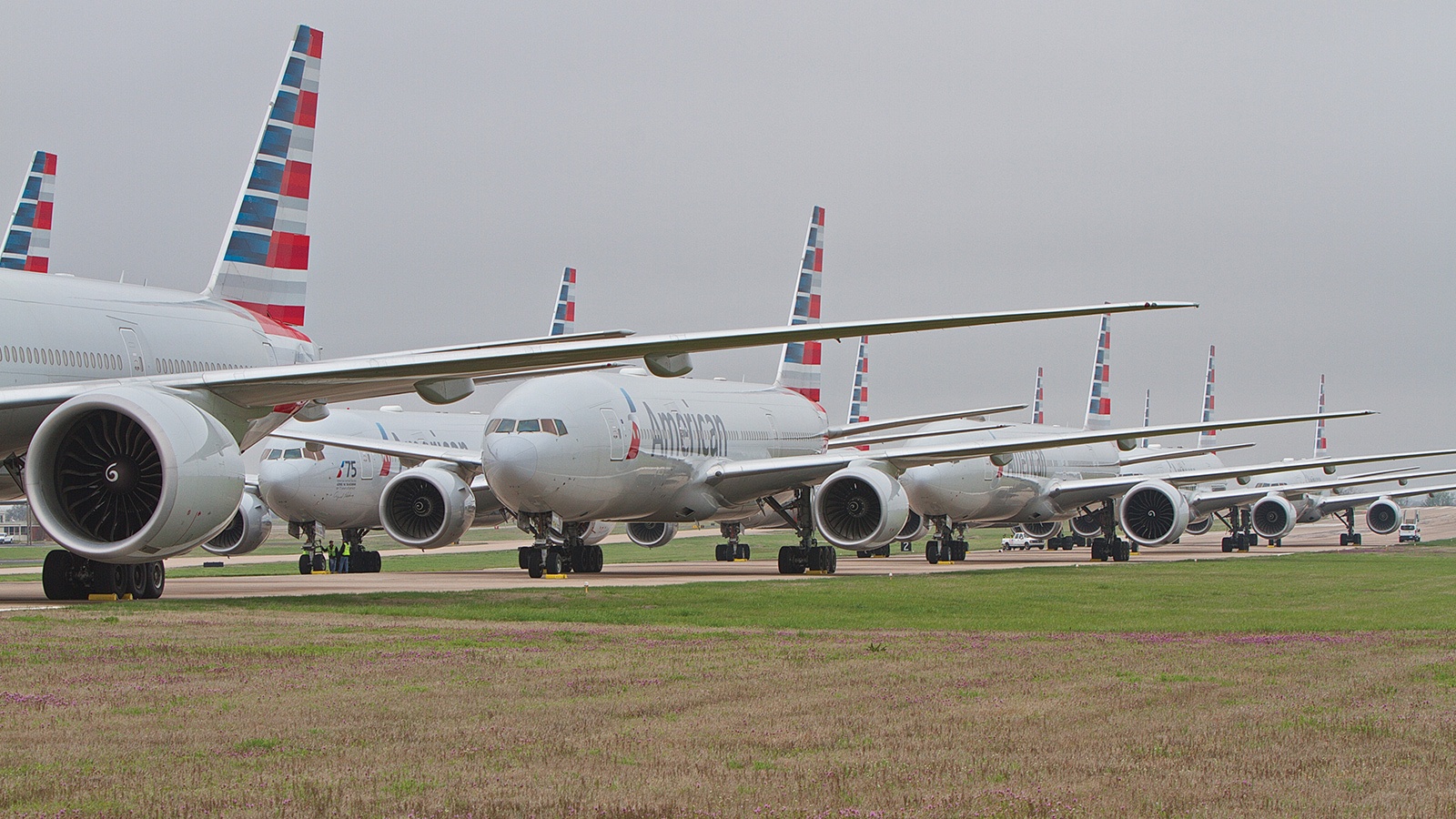 Several commercial airplanes with American Airlines livery are lined up on a runway, positioned closely together under a cloudy sky.