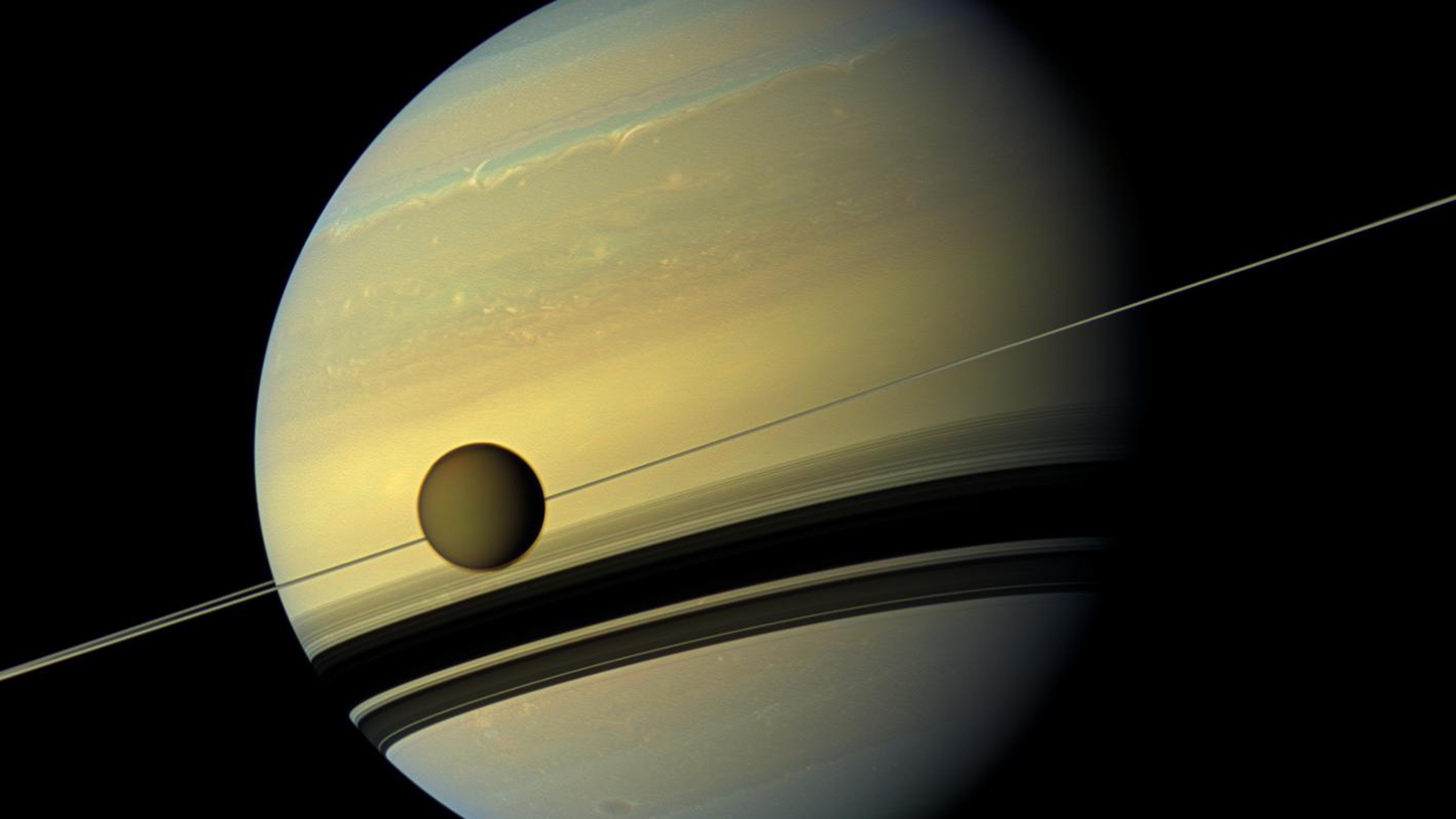 Image of Saturn with its rings, with one of its moons, likely Titan, positioned in front of the planet. The background is the dark expanse of space.