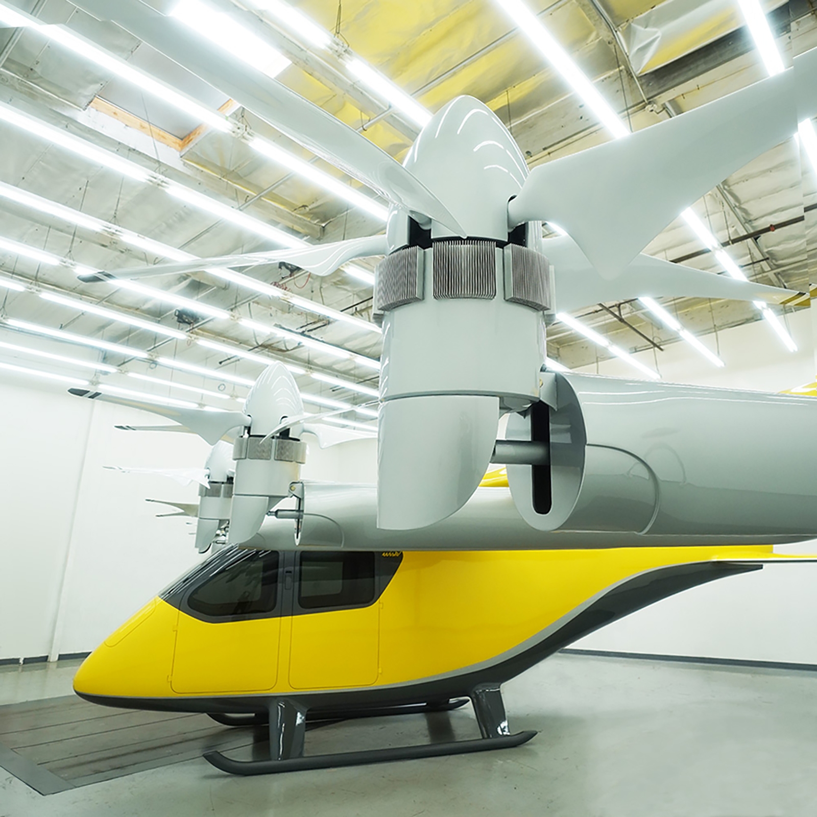 A futuristic yellow and gray vertical takeoff and landing aircraft with large propellers is displayed in a well-lit indoor facility.