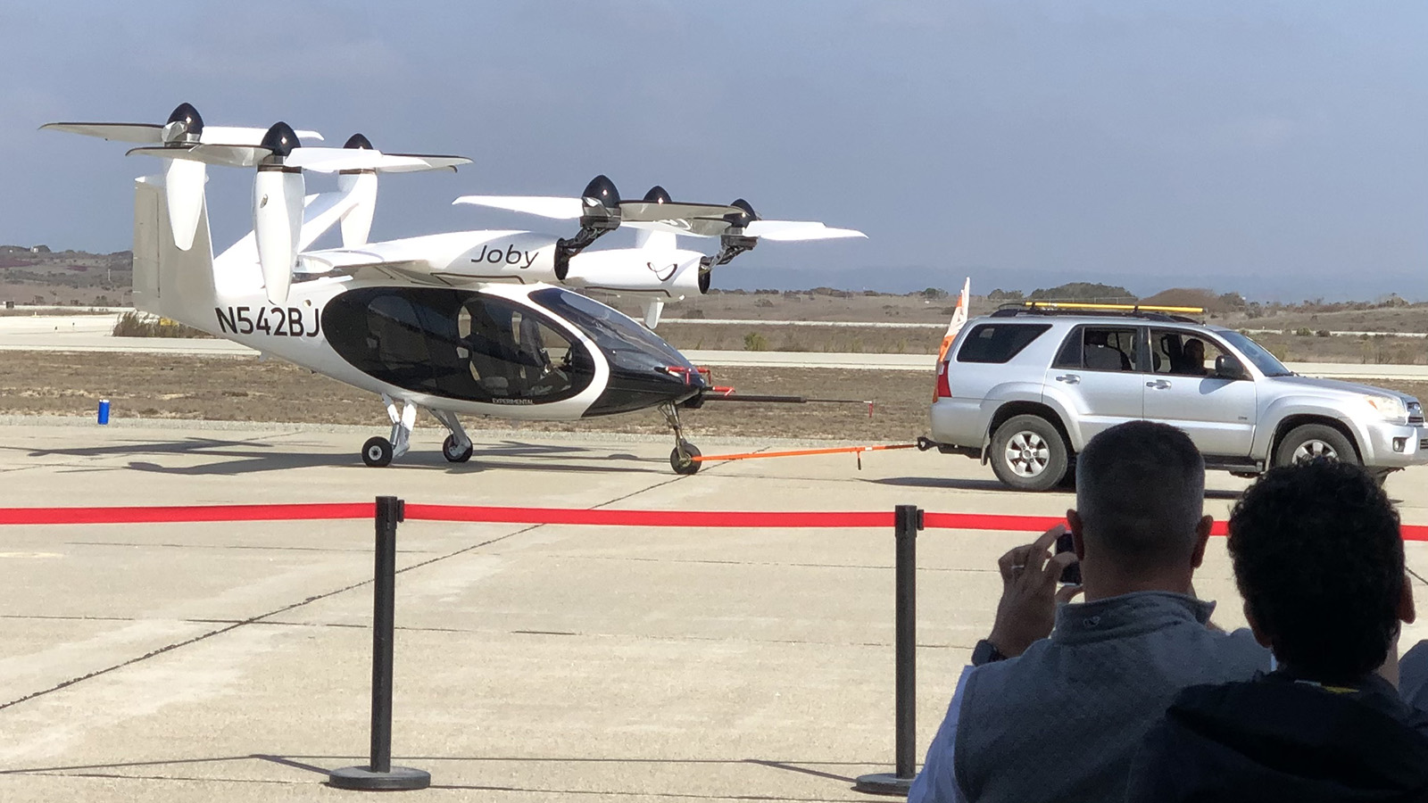 A small white and black aircraft with the tail number N542BJ is being towed by a white SUV on an airport tarmac. Several people in the foreground are observing and taking photos.