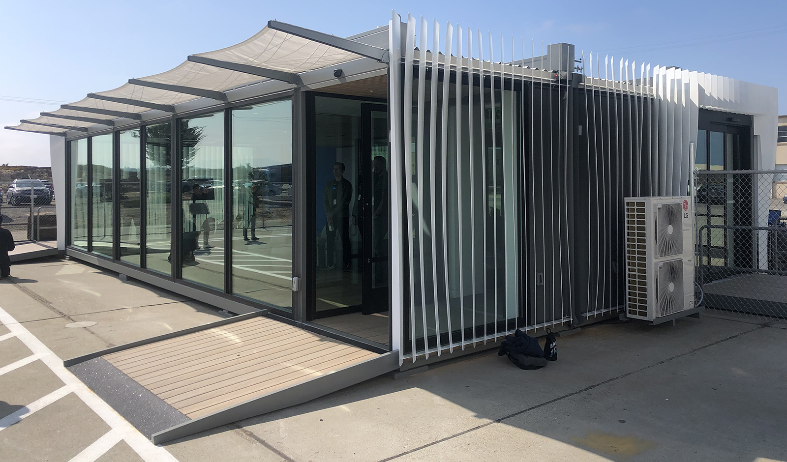 Modern, single-story building with large glass windows, an overhang for shade, and a small wooden ramp leading to the entrance. An outdoor air conditioning unit is visible on the right side.