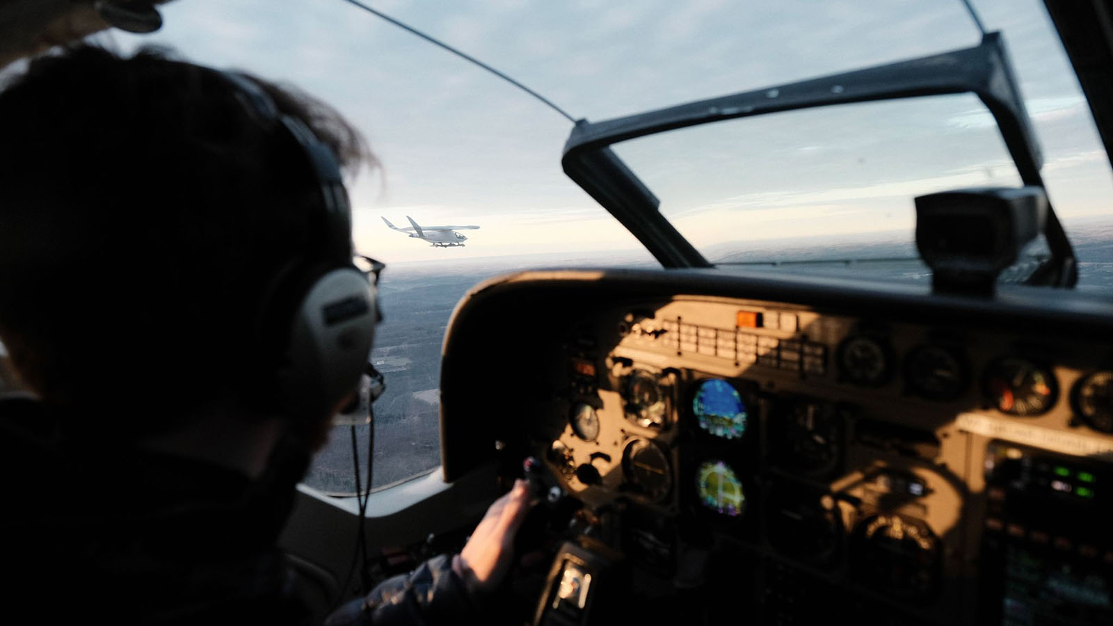 View from inside an airplane cockpit with a pilot holding the controls, looking at another airplane flying ahead in the distance against a backdrop of a cloudy sky.