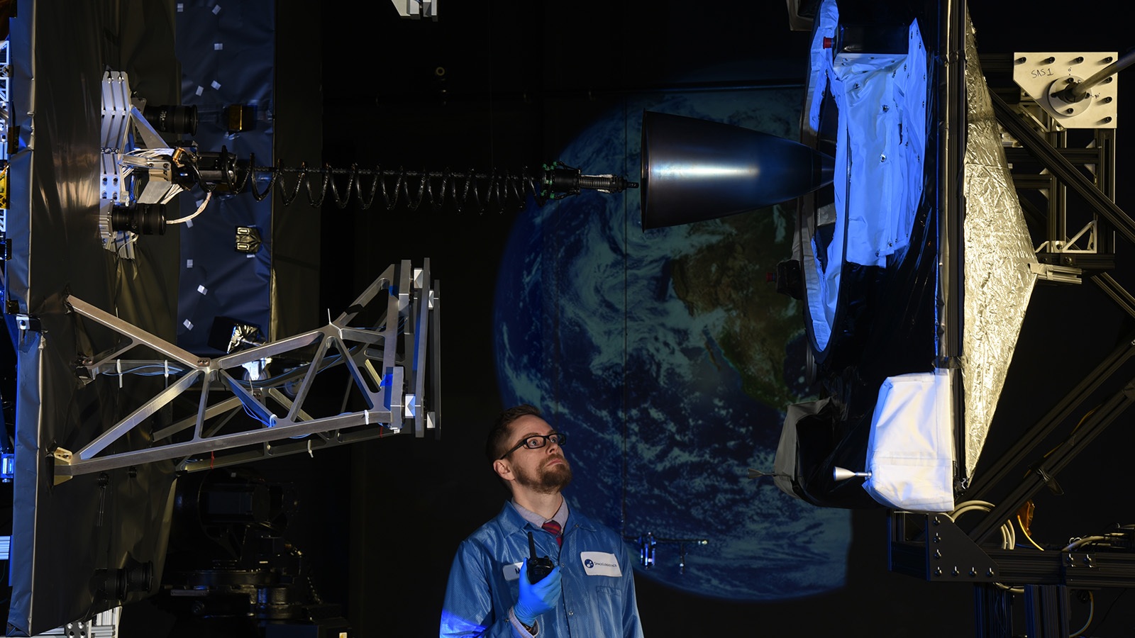 A man in a blue lab coat inspects space equipment inside a lab, with a large image of Earth projected in the background.