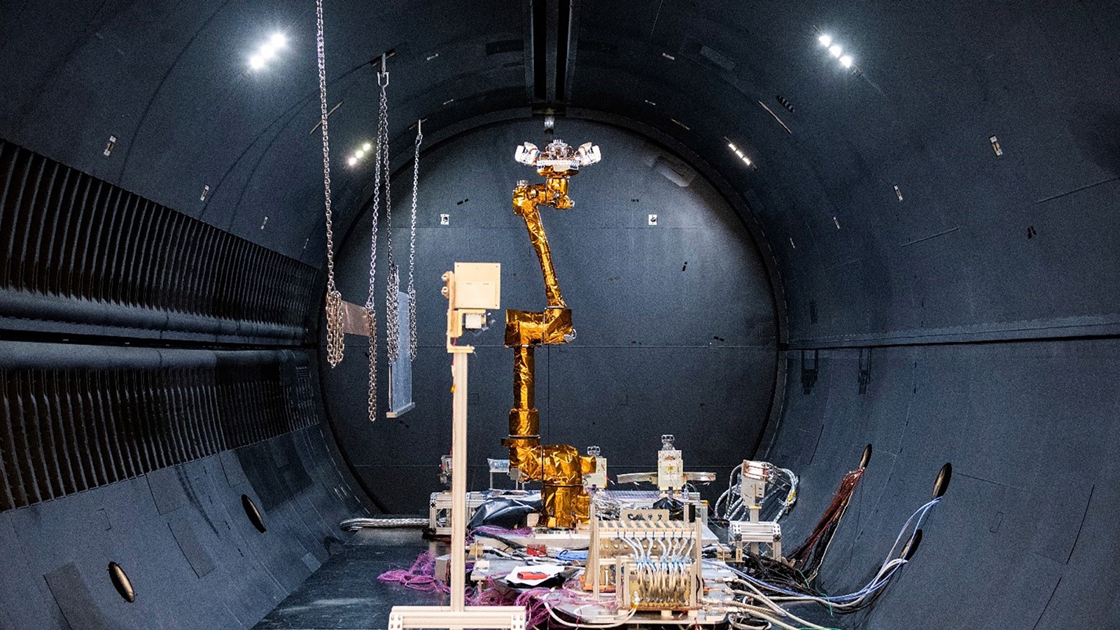 A robotic arm manipulator inside a large, dimly-lit chamber with various cables and equipment attached, suggesting a scientific or industrial testing environment.