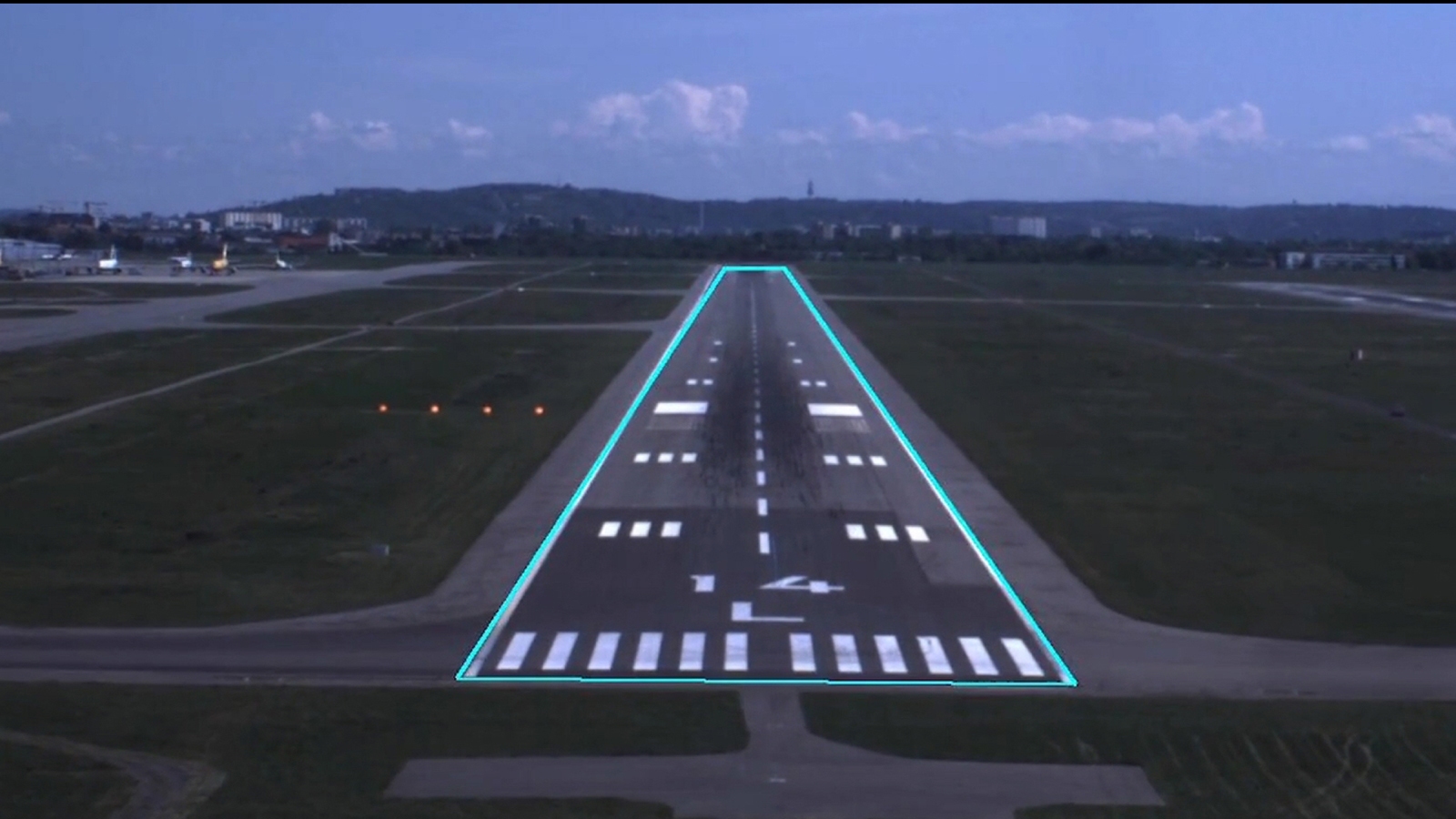 A view of an illuminated airport runway during nighttime, marked with alternating white stripes and flanked by green grass on both sides. The sky is dark with faint clouds in the background.