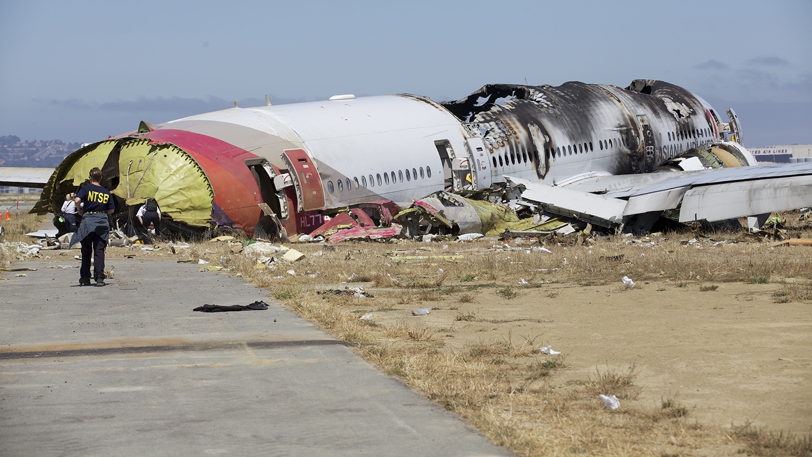 The image shows the wreckage of a burned-out airplane on a clear day, with debris scattered around and an investigator from the NTSB examining the scene.