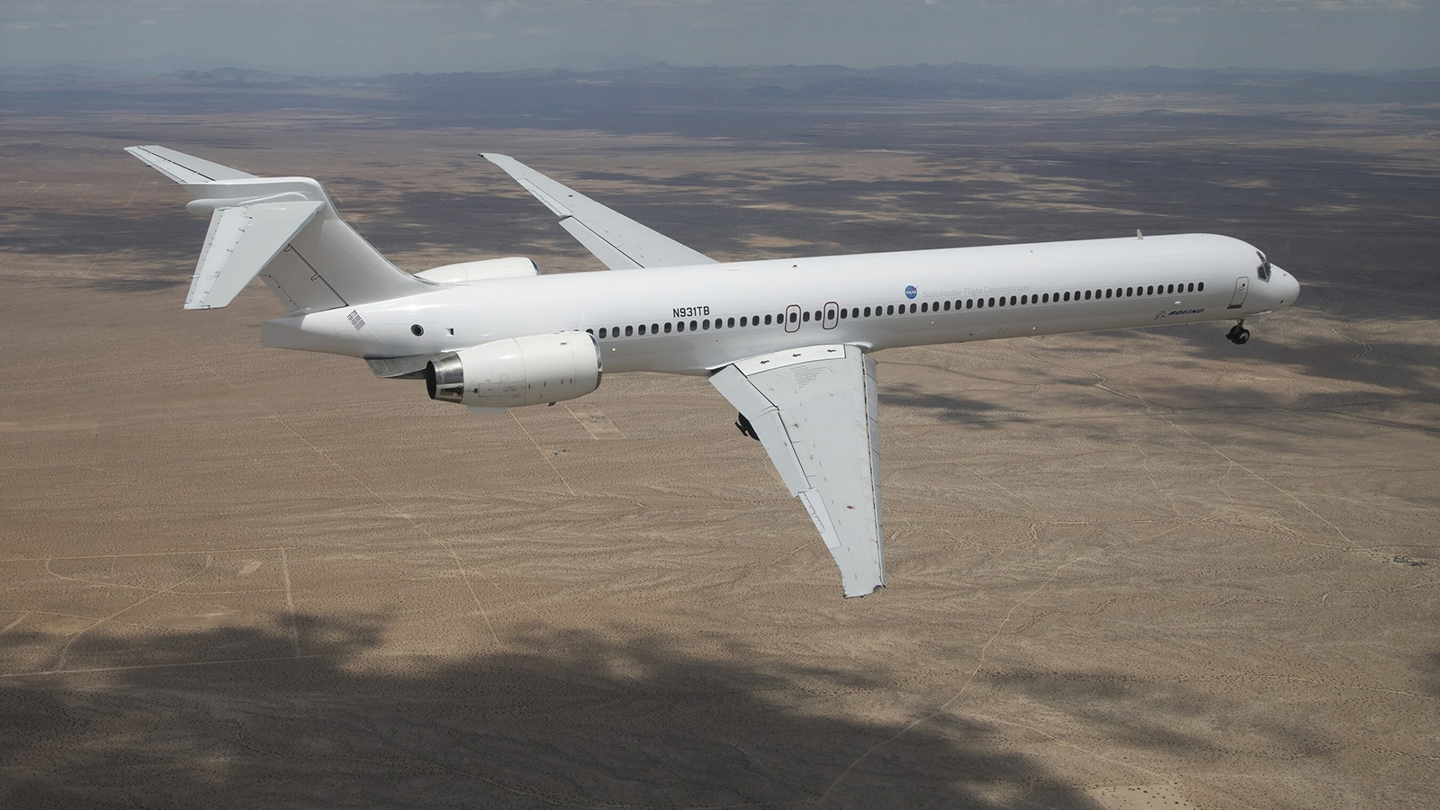 A white jet airplane flies over a barren desert landscape during the daytime.