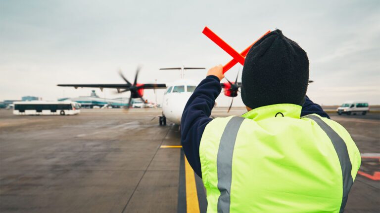 February 2024 AIAA Bulletin A ground crew worker in a high-visibility jacket guides an approaching propeller aircraft on an airport tarmac using red directional wands.