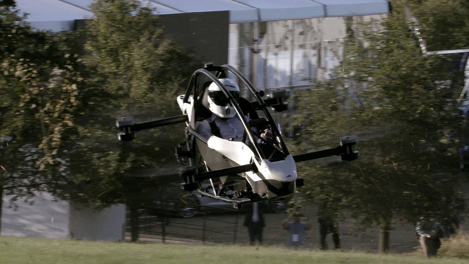 A person is piloting a small, open-cockpit, electric aircraft with multiple rotors, flying close to the ground in an outdoor area near trees and a building structure.