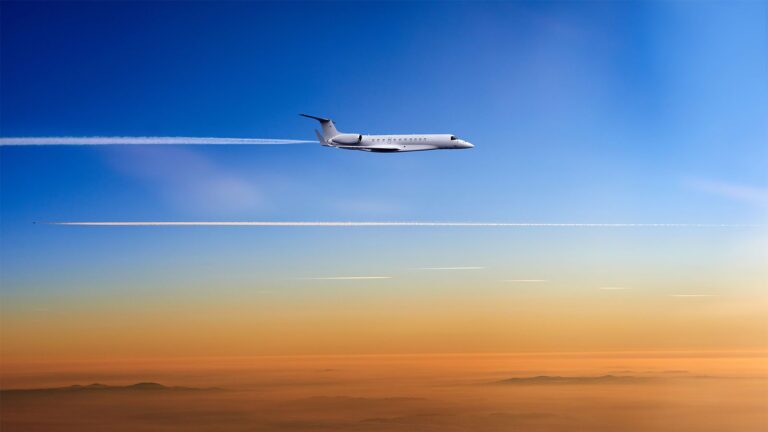 A jet airplane flies at high altitude leaving contrails behind, against a backdrop of a clear blue sky and a gradient of warm colors near the horizon.