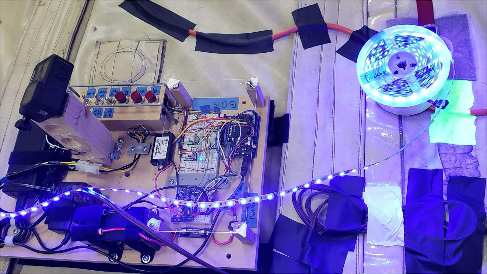 A wooden board with an assembly of electronic components and wiring, including an Arduino, power supply, buttons, and a connected LED strip that coils beside the board, emitting blue light.