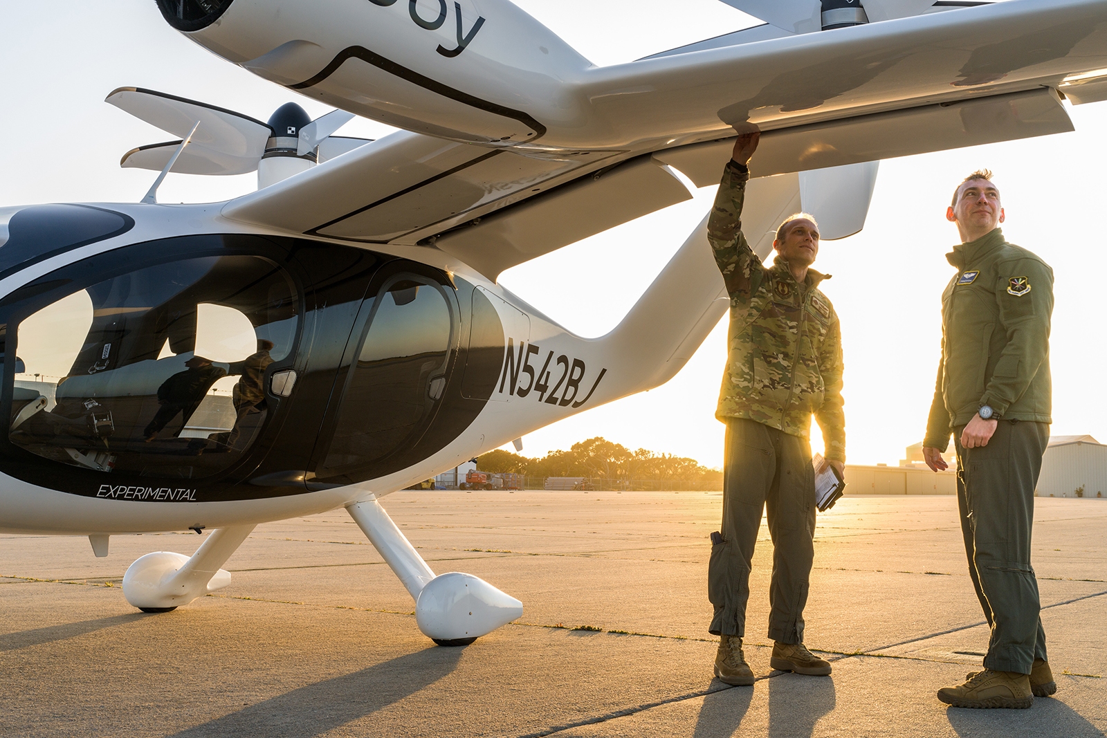 Two people in military attire examine a white aircraft on an airfield at sunset. One person points to the wing while the other looks on.