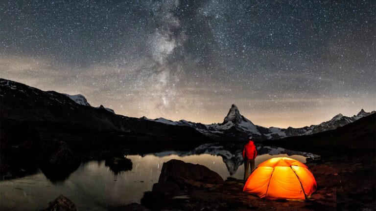 A person stands near an illuminated tent beside a lake, looking at the starry night sky with a prominent mountain peak in the background.