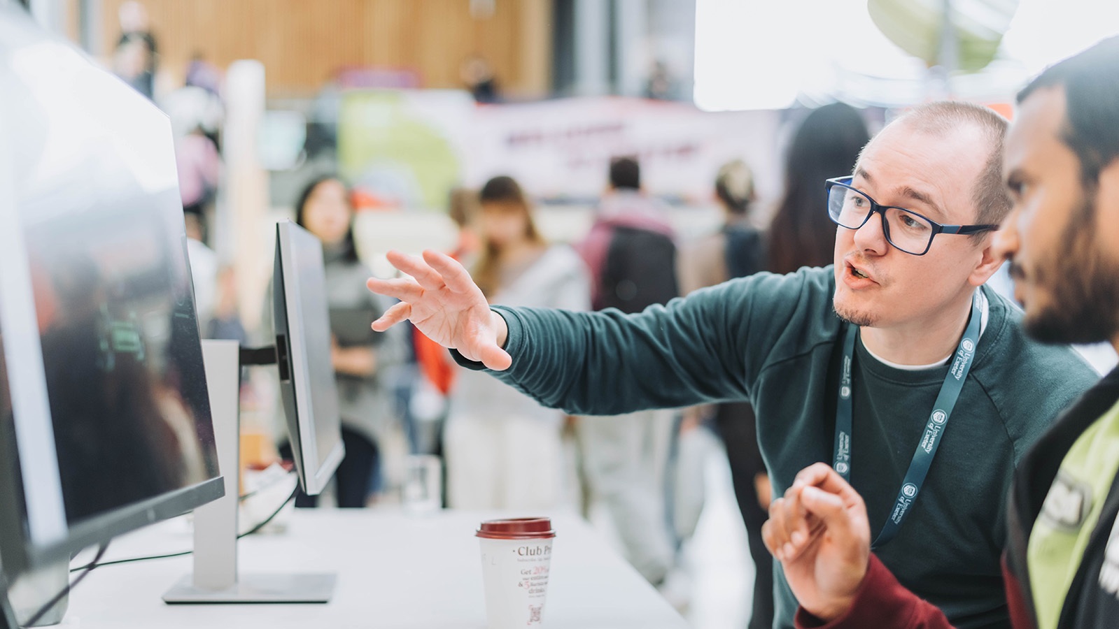 Two people discussing in front of computer screens. One person is gesturing while the other listens. There's a coffee cup on the table.