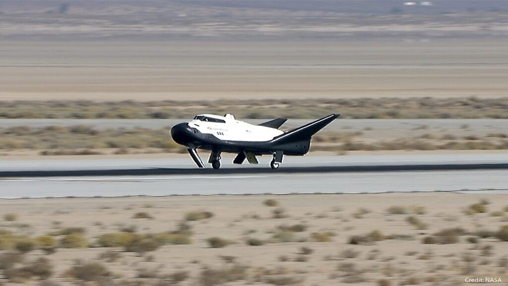 A small futuristic-looking spacecraft with a white body and black wings is landing on a runway in a desert landscape. Credit: NASA.