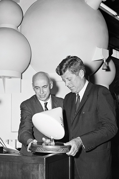 Two men in suits examine a modern light fixture on a wooden base. Large round light fixtures hang in the background.